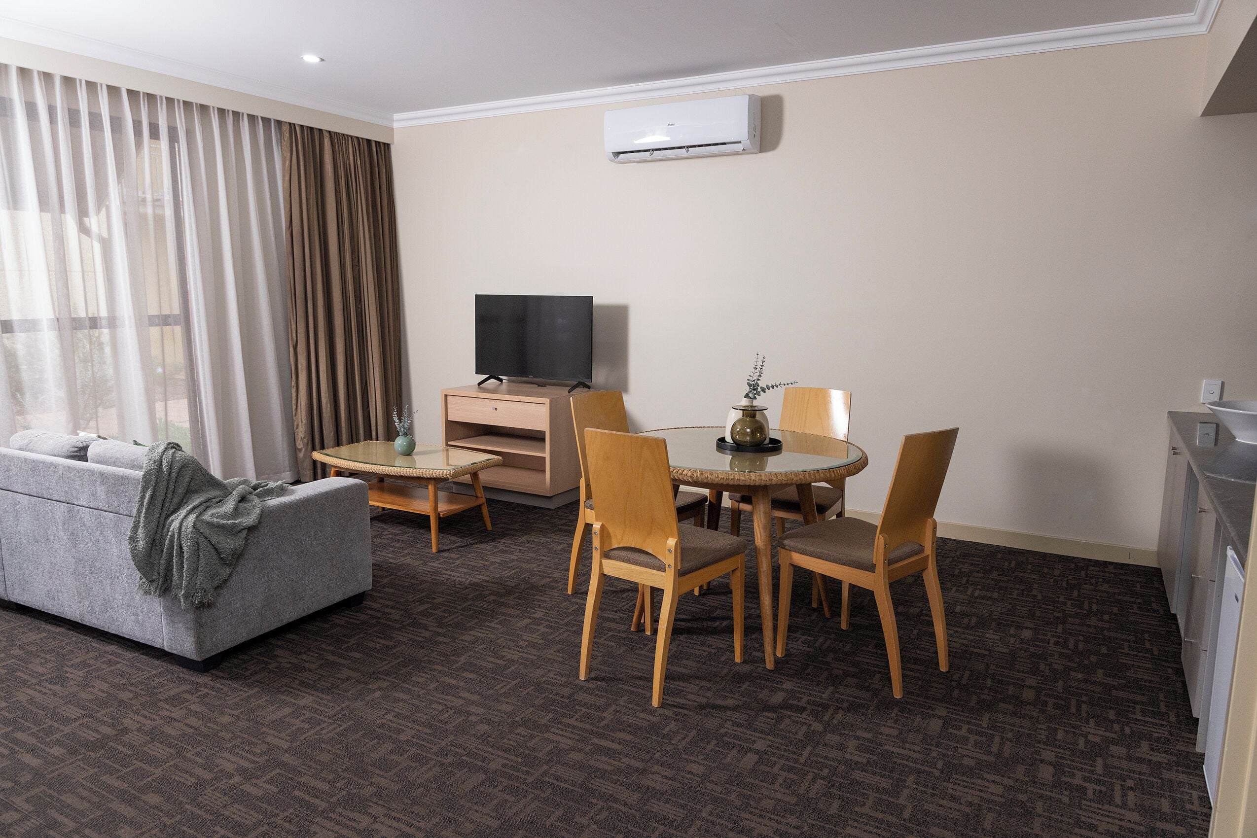 A hotel room with a sofa, a TV, a dining table with chairs, and an air conditioning unit.