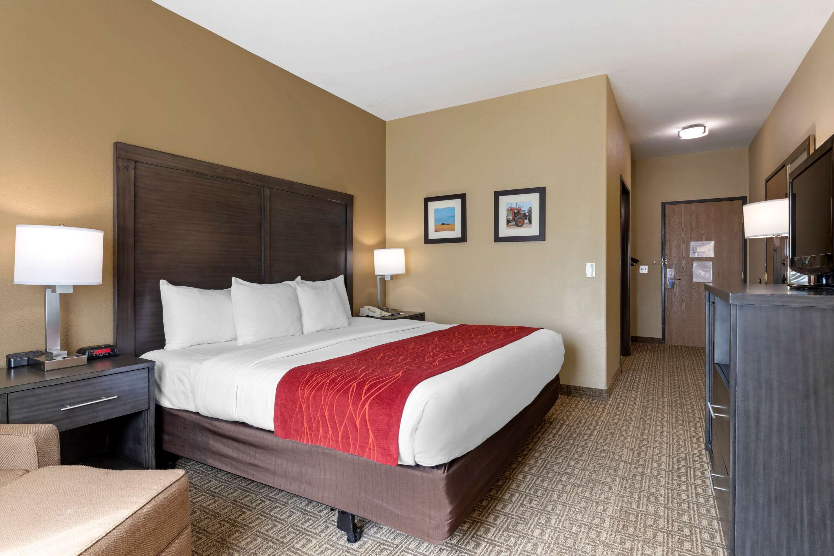 A hotel room with a large bed, a desk with a lamp, a chair, a dresser, and a door leading to another room.