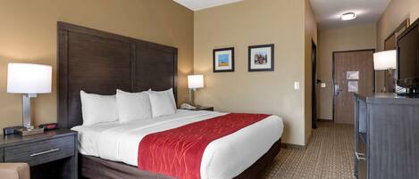 A hotel room with a large bed, a desk with a lamp, a chair, a dresser, and a door leading to another room.