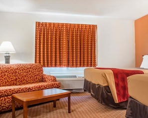 A hotel room with a patterned sofa, a wooden table, a bed with a red blanket, and an air conditioning unit. - Econo Lodge Cuba I-86 (Cuba)