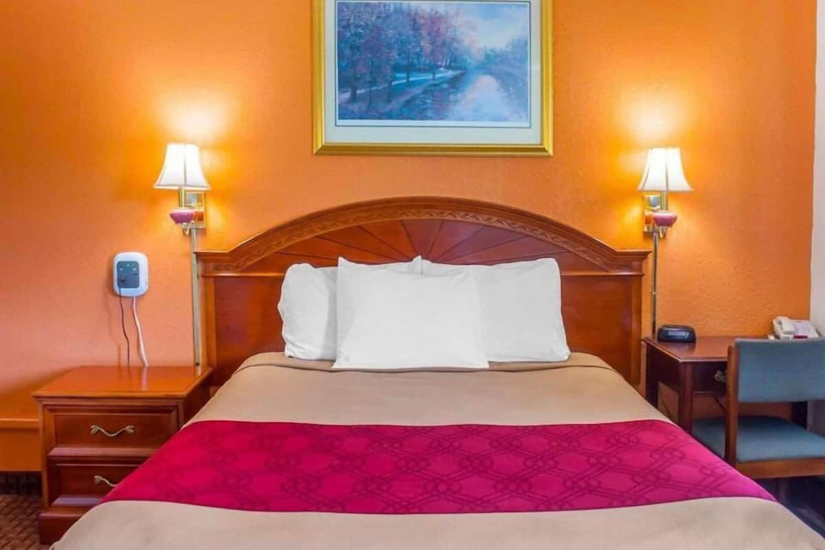 A hotel room with a bed, two bedside lamps, a nightstand, a desk, and a painting on the wall.