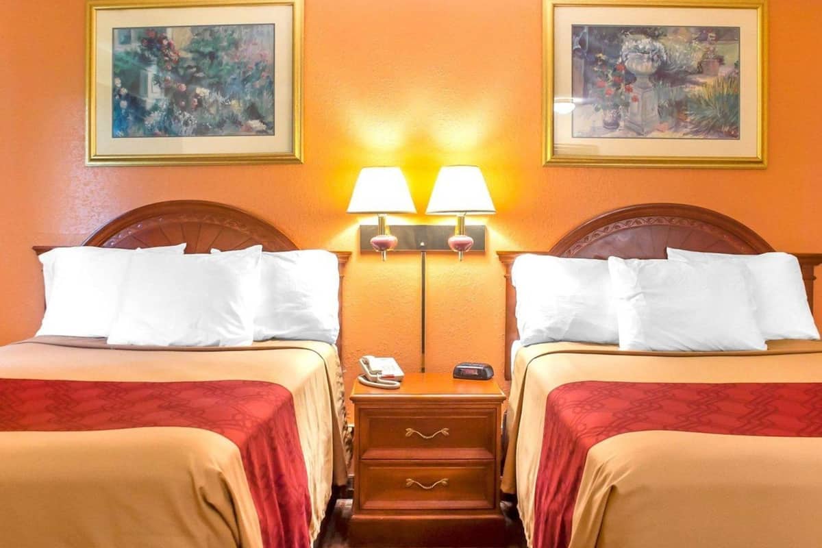 A hotel room with two beds, a nightstand with a phone and remote, and two wall-mounted lamps.