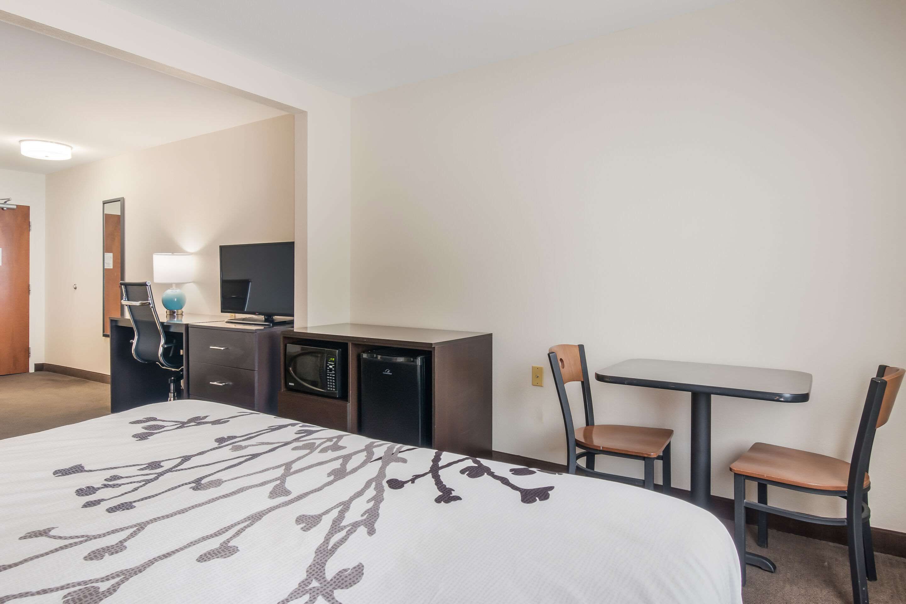 Quarto de hotel com uma cama, uma escrivaninha com computador e luminária, um micro-ondas e duas cadeiras.