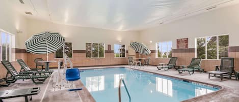 An indoor swimming pool with lounge chairs and umbrellas, surrounded by windows and a tiled wall.