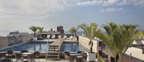 A rooftop pool area with lounge chairs and tables, a small waterfall feature, and a view of the ocean.