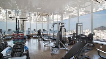Un gimnasio con diversos equipos de ejercicio, ventanas amplias y vistas a un paisaje urbano.