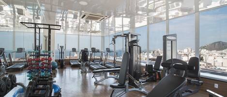 Un gimnasio con diversos equipos de ejercicio, ventanas amplias y vistas a un paisaje urbano.