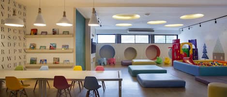 Una habitación luminosa con una mesa larga, sillas coloridas y una zona de juegos con un tobogán y piscina de pelotas.