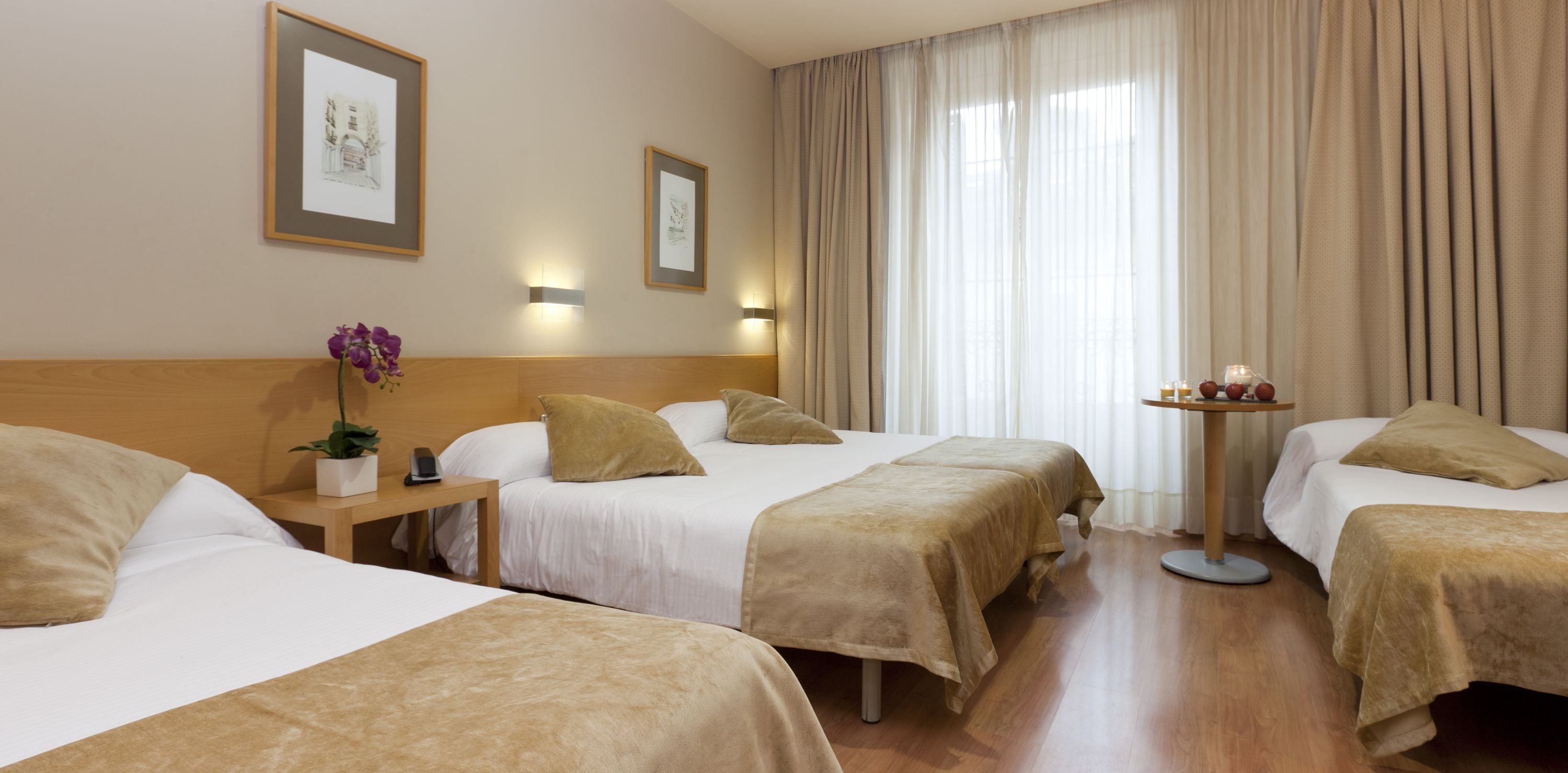 A hotel room with three beds, a bedside table with a lamp, a small table with candles, and a window with curtains.
