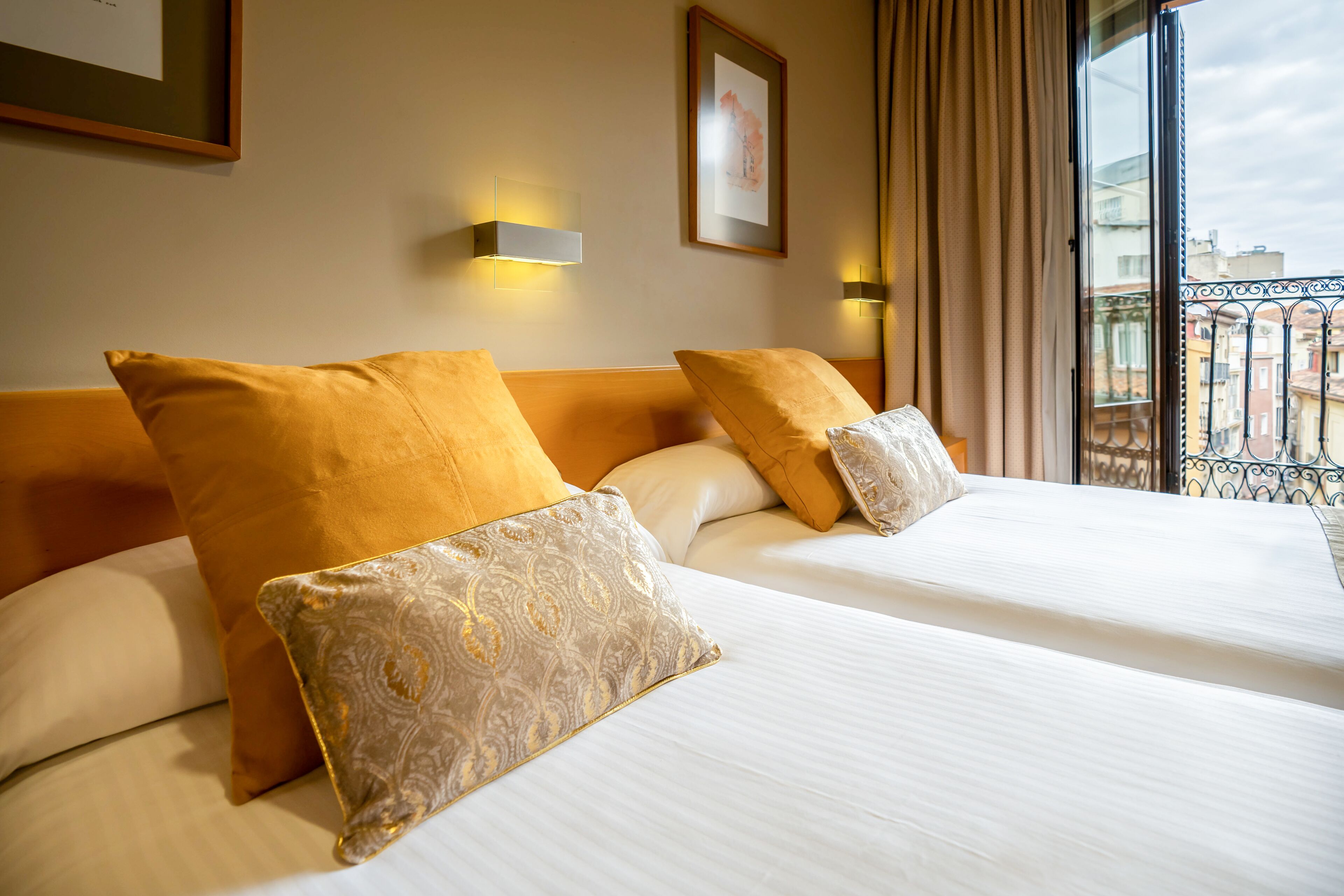 A hotel room with two beds, each with white linens and decorative pillows. There are wall-mounted light fixtures and a framed picture on the wall.