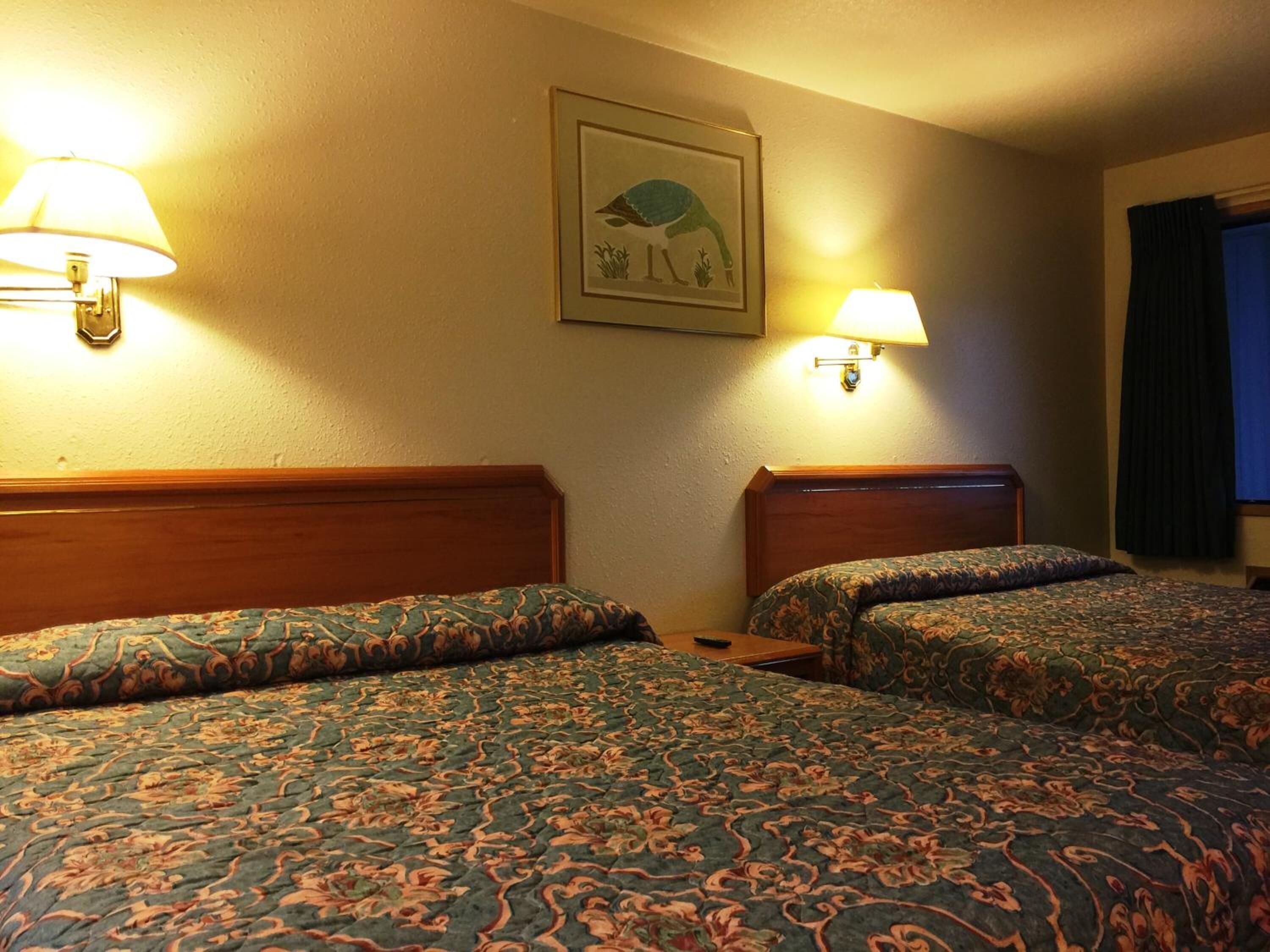 A hotel room with two beds, each with a patterned bedspread and headboard, a framed picture on the wall, and two wall-mounted lamps.