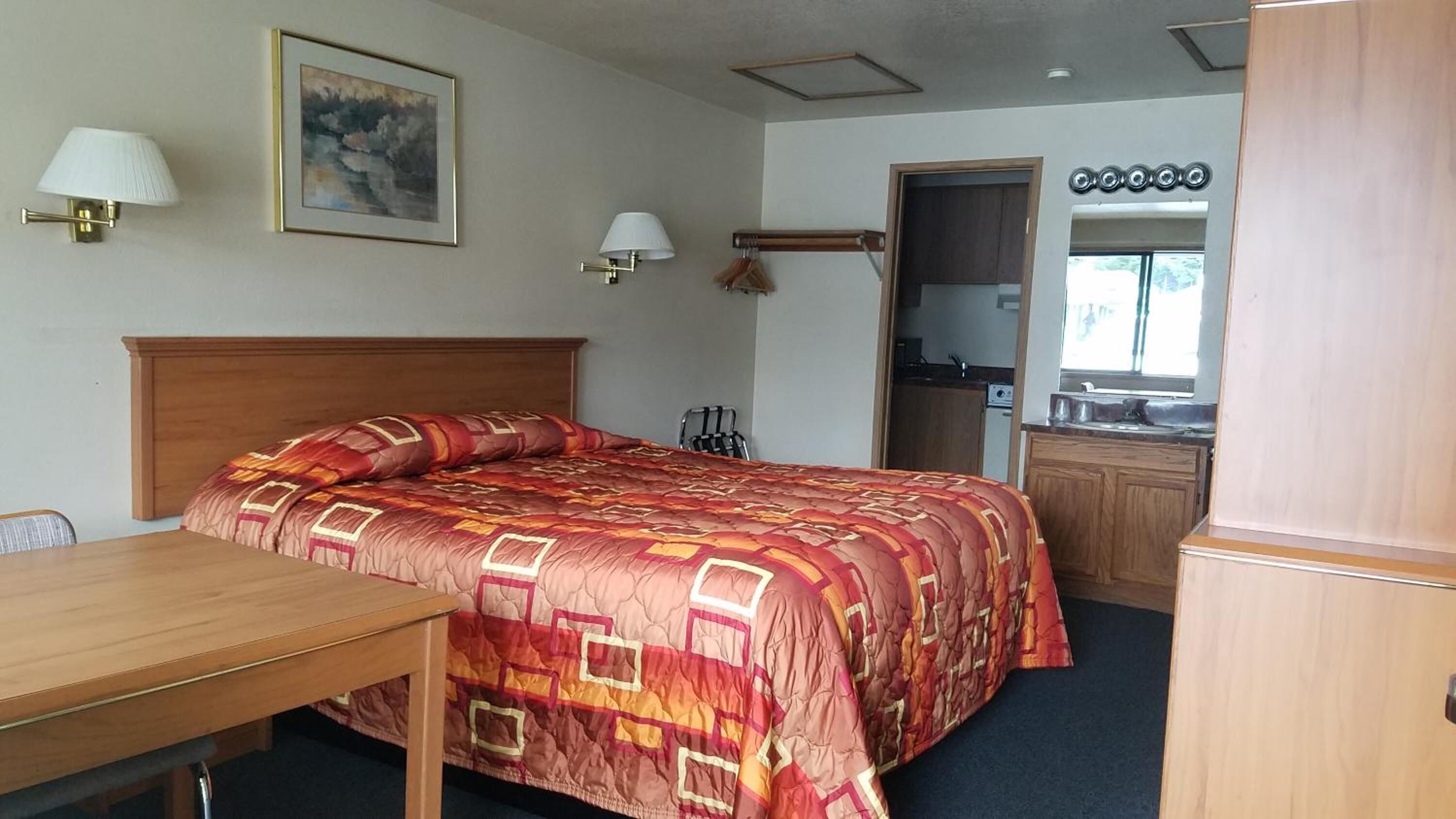 Habitació d'hotel amb un llit de fusta, un escriptori, una cadira, un quadre a la paret i una cuina visible a través d'una porta oberta.