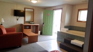 A hotel room with a sofa, a small desk, a television, a green door, a bathtub, and a window.