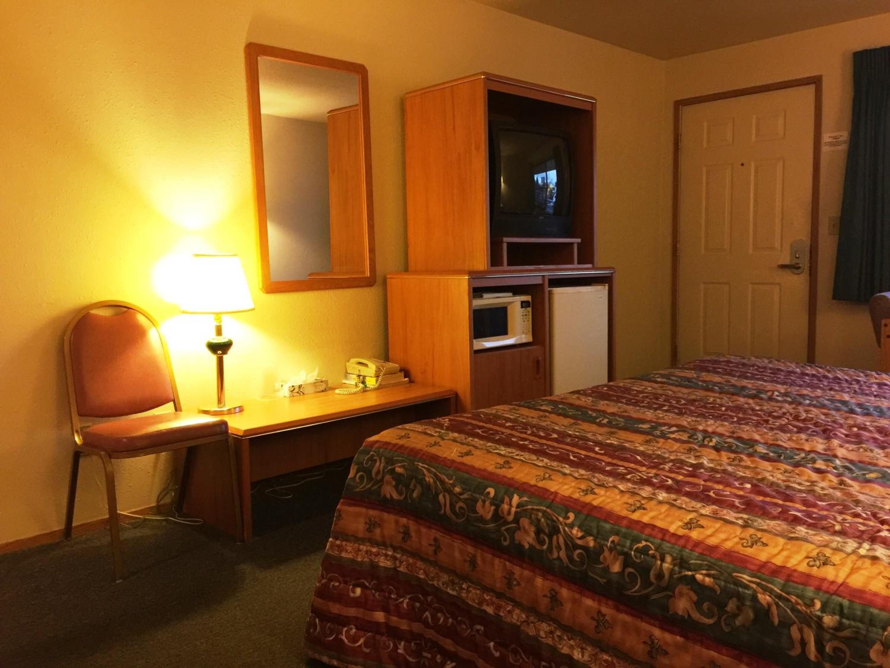 A hotel room with a bed, a desk with a lamp, a microwave, a refrigerator, a mirror, and a door.