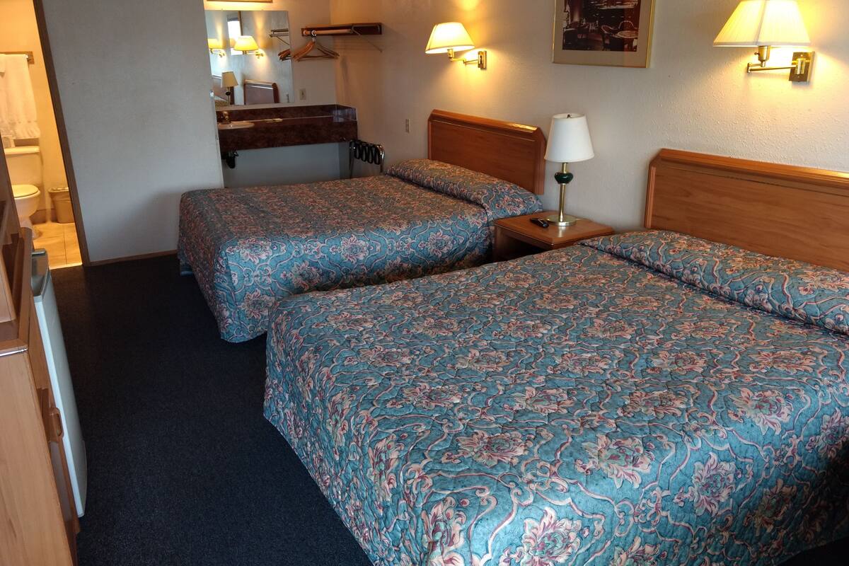 A hotel room with two beds, a wooden headboard, a bedside table with a lamp, a mirror, a towel rack, and a small bathroom with a toilet.