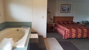 A bathroom with a bathtub, a sink, and a towel. There is a bed with a red patterned bedspread and a nightstand with a lamp.