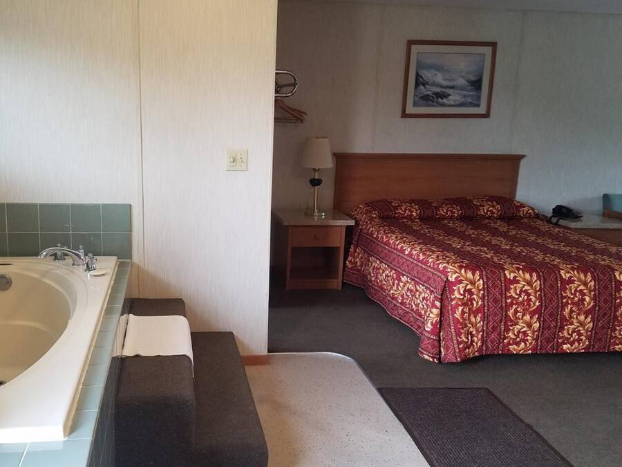 A bathroom with a bathtub, a sink, and a towel. There is a bed with a red patterned bedspread and a nightstand with a lamp.