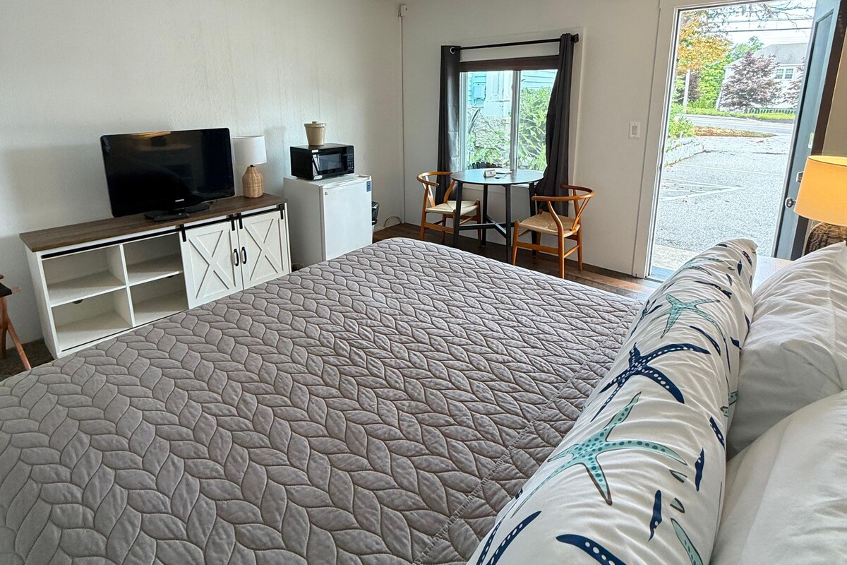 A hotel room with a large bed, a desk with a TV and microwave, a small table with chairs, and a window with a view of the outside.
