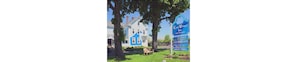 A large white building with a blue roof, surrounded by trees and a signboard advertising a hotel with a vacancy. - Coast Village Inn & Cottages (Wells)