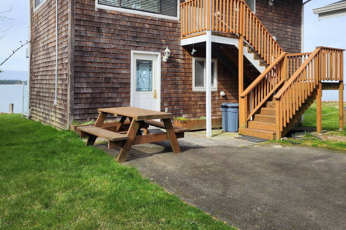 A two-story wooden house with a balcony, a wooden staircase, and a picnic table.