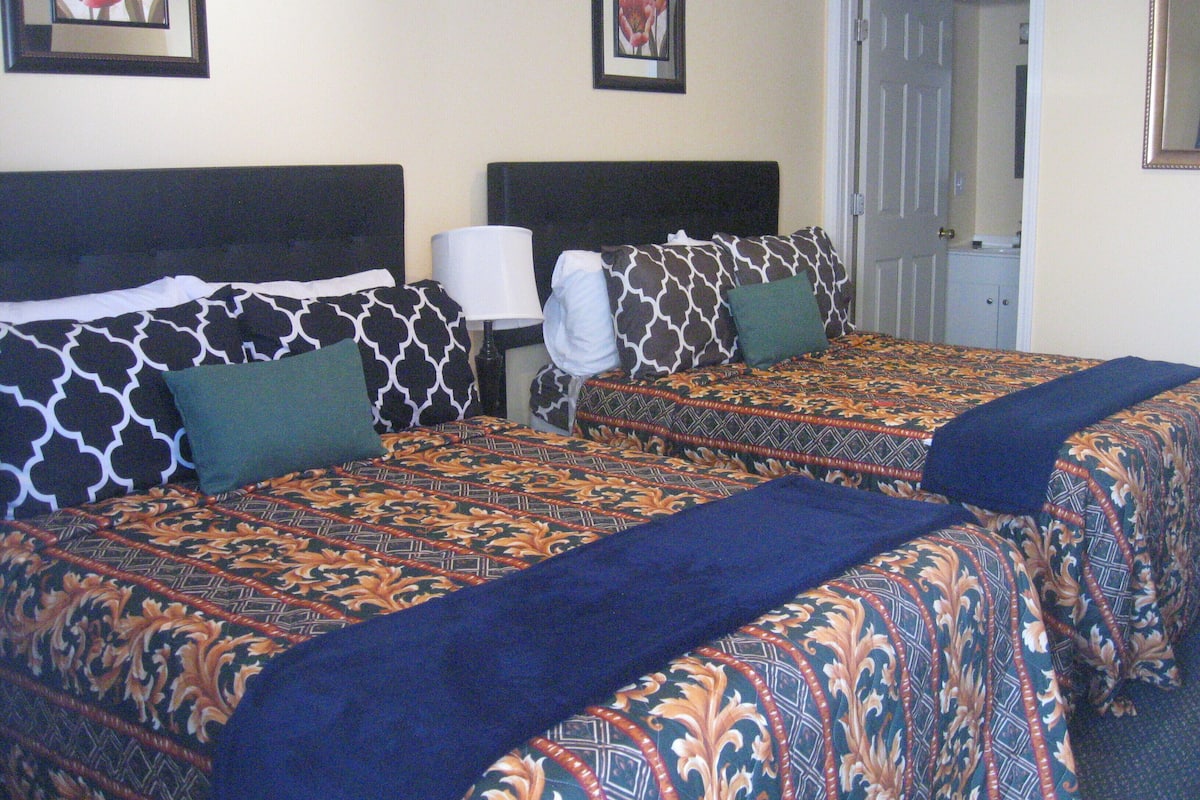 A hotel room with two beds, each with patterned bedding and pillows. There is a nightstand with a lamp between the beds.