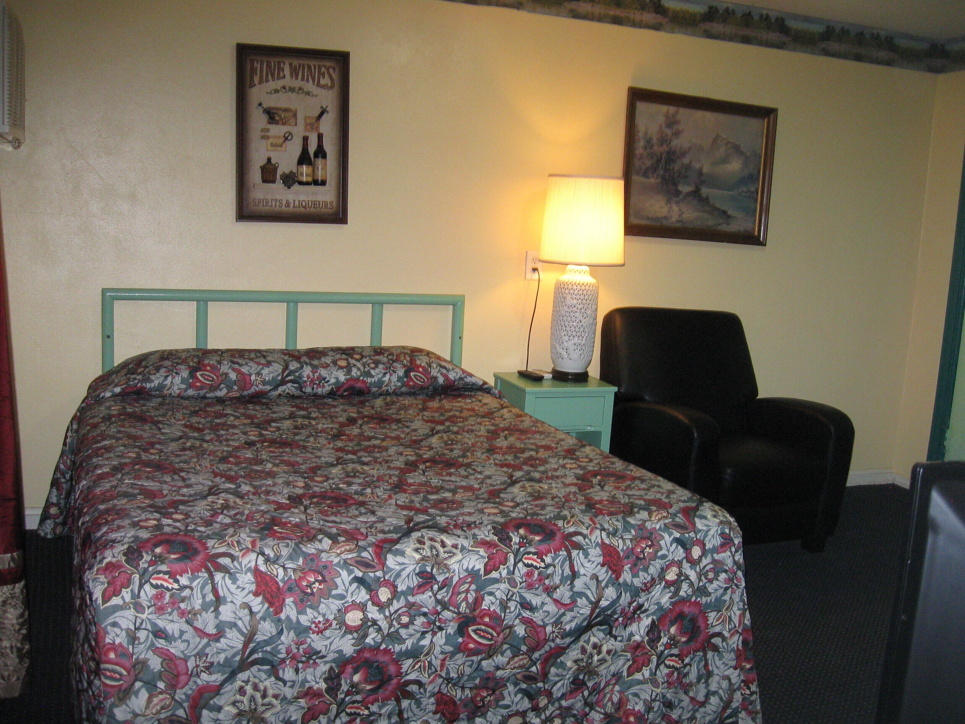A hotel room with a floral bedspread, a framed poster on the wall, a lamp, and a chair.