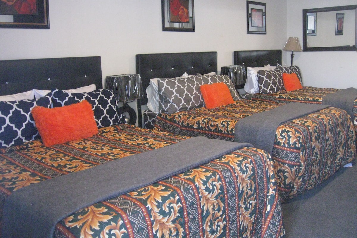 A hotel room with three beds, each with patterned bedding and orange pillows. There are framed pictures on the walls and a lamp on the nightstand.
