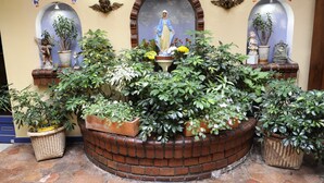 Uma estátua de figura religiosa cercada por vasos com plantas e flores, com elementos decorativos nas paredes.