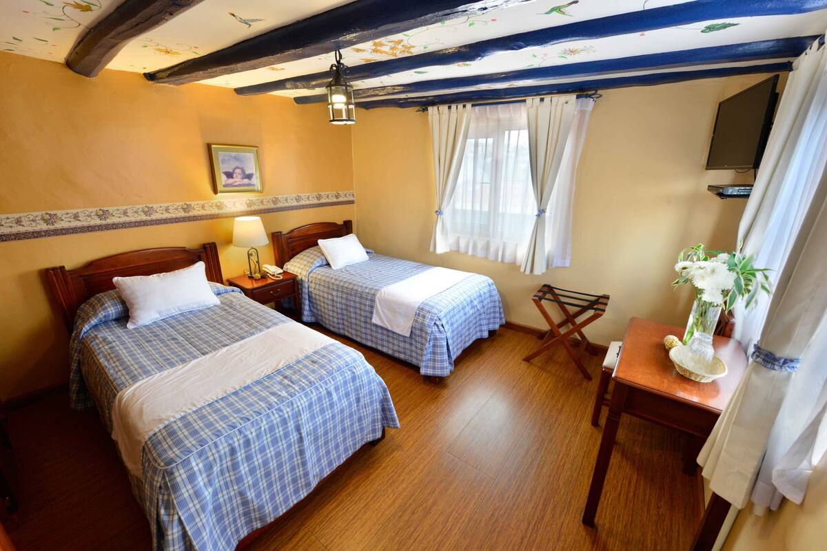 Habitación de hotel con dos camas, suelo de madera, una mesita con un jarrón de flores y techo con vigas a la vista.