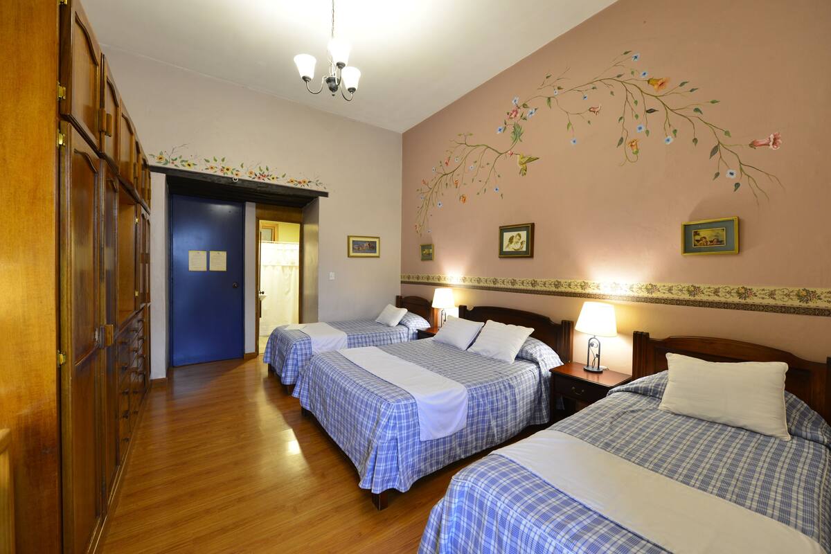 Habitación de hotel con tres camas, suelo de madera, un mural floral en la pared y una araña.