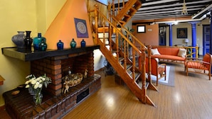 Escada de madeira que leva a um andar superior, lareira de tijolos com objetos decorativos e um espaço de estar aconchegante com móveis laranja.