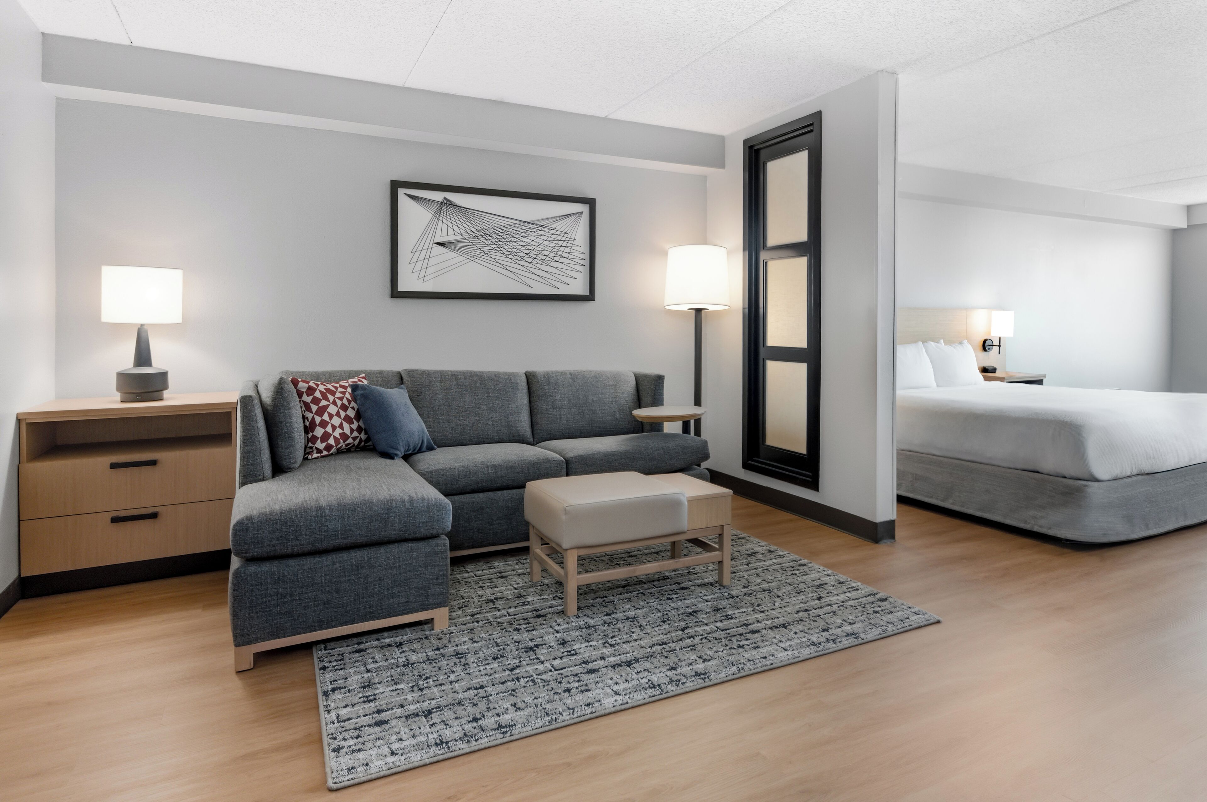 A modern hotel room with a grey sectional sofa, a small ottoman, a grey rug, a bedside table with a lamp, and a bed with white linens.