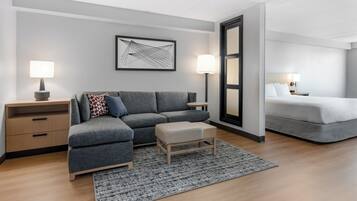 A modern hotel room with a grey sectional sofa, a small ottoman, a grey rug, a bedside table with a lamp, and a bed with white linens.