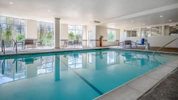 An indoor swimming pool with clear glass walls, multiple seating areas, and a view of trees outside.
