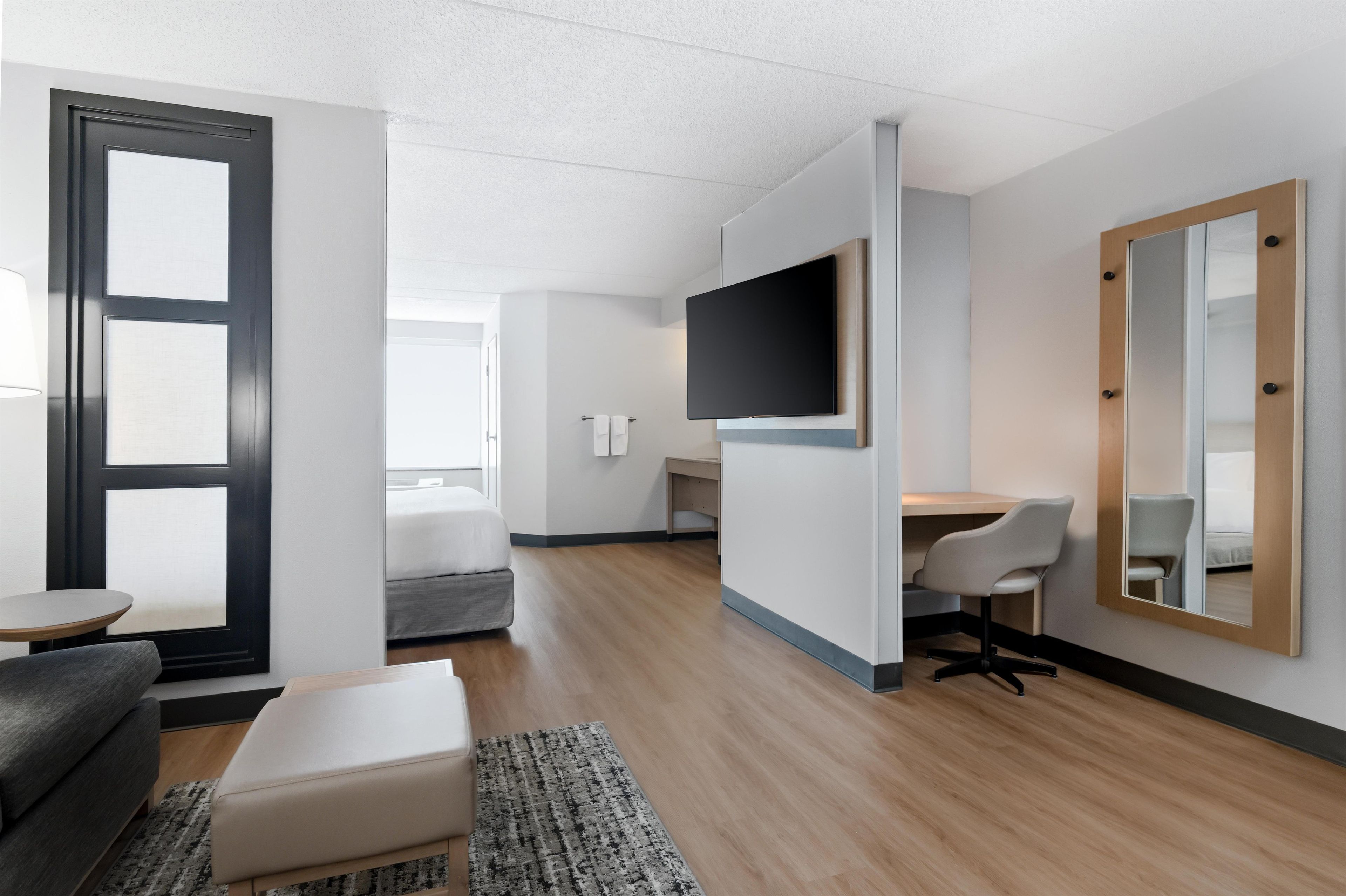 A modern hotel room with a bed, a desk, a chair, a TV, and a large mirror.