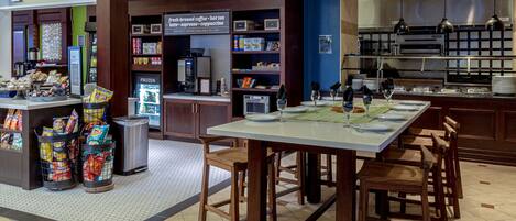 Una cafetería con mostrador que exhibe aperitivos y bebidas, zona de estar con mesas y sillas, y vitrina refrigerada.