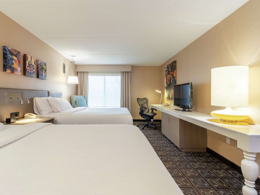Habitación de hotel con dos camas, un escritorio con televisor, una silla y obras de arte en las paredes.