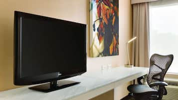 A modern hotel room with a large flat-screen TV on a marble countertop, a black office chair, and a painting on the wall.