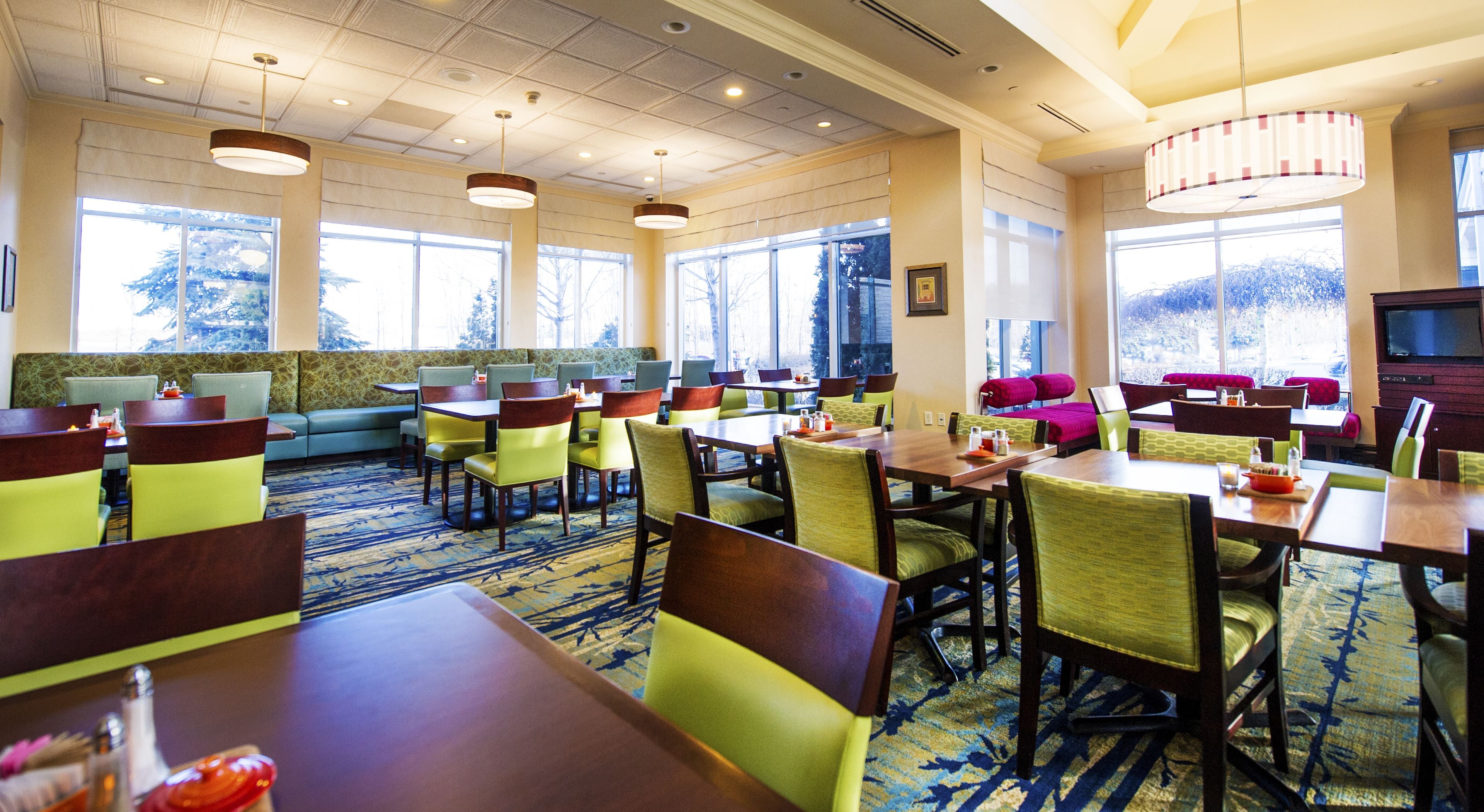 A hotel dining area with multiple tables and chairs, large windows, and a carpeted floor.