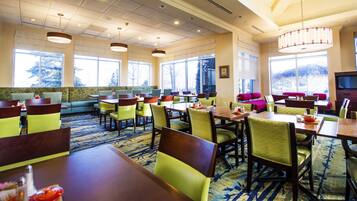 A hotel dining area with multiple tables and chairs, large windows, and a carpeted floor.