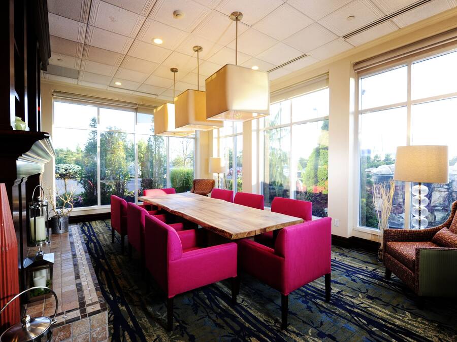 Un comedor de hotel con una gran mesa de madera rodeada de sillas rosa brillante, ventanas amplias que ofrecen vistas al exterior e iluminación colgante moderna.