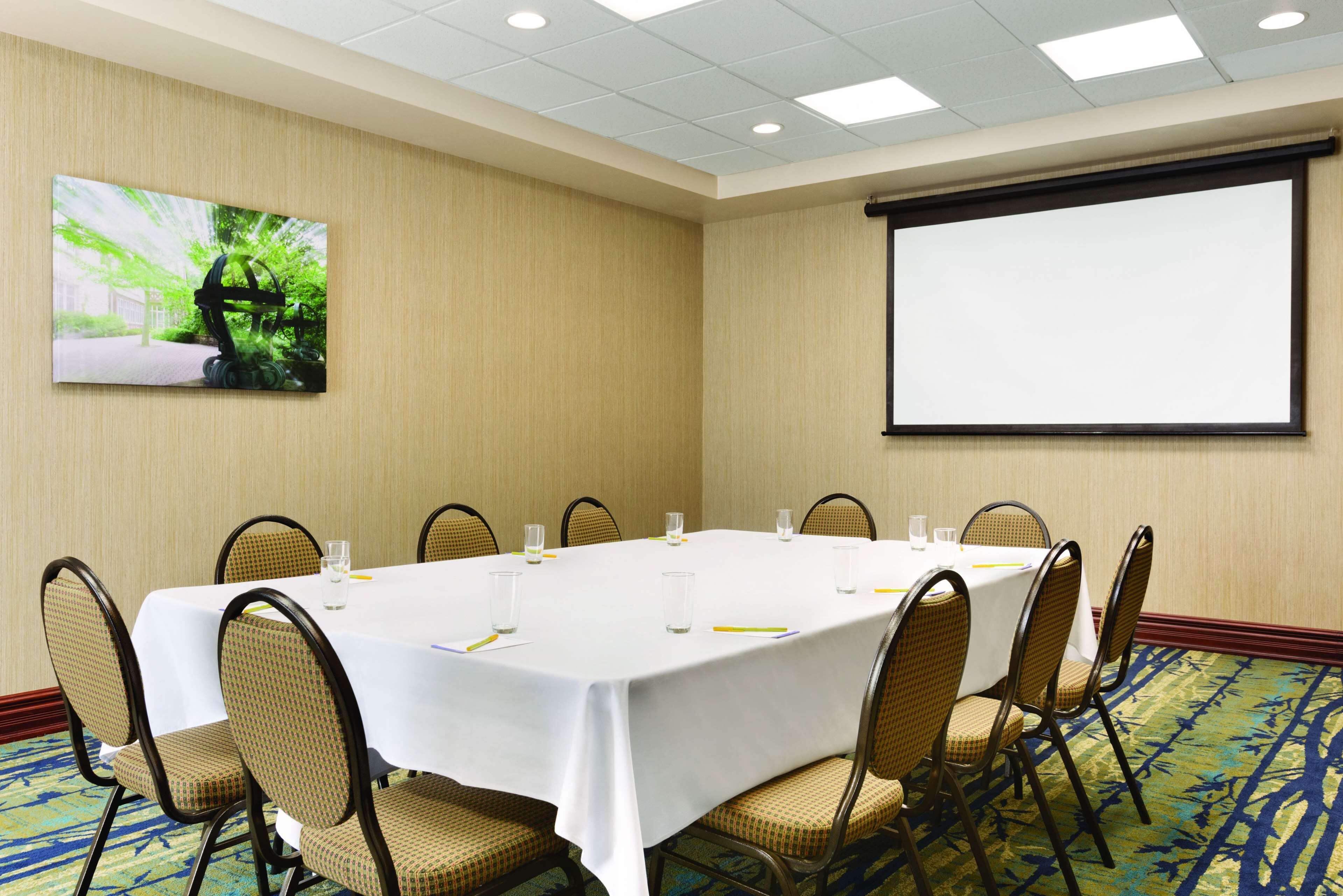 Un salón de conferencias con una mesa blanca, sillas, una pantalla de proyector y un cuadro en la pared.