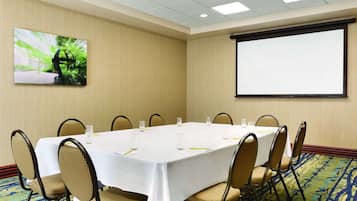 Un salón de conferencias con una mesa blanca, sillas, una pantalla de proyector y un cuadro en la pared.