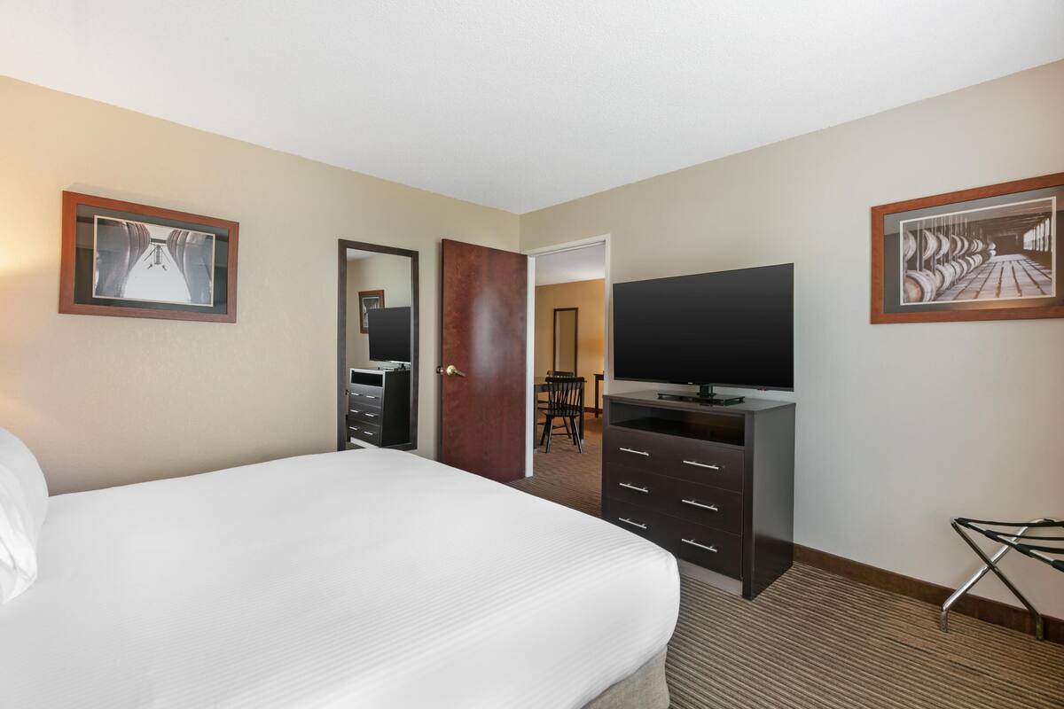 A hotel room with a bed, a TV, a dresser, and a door leading to a dining area.