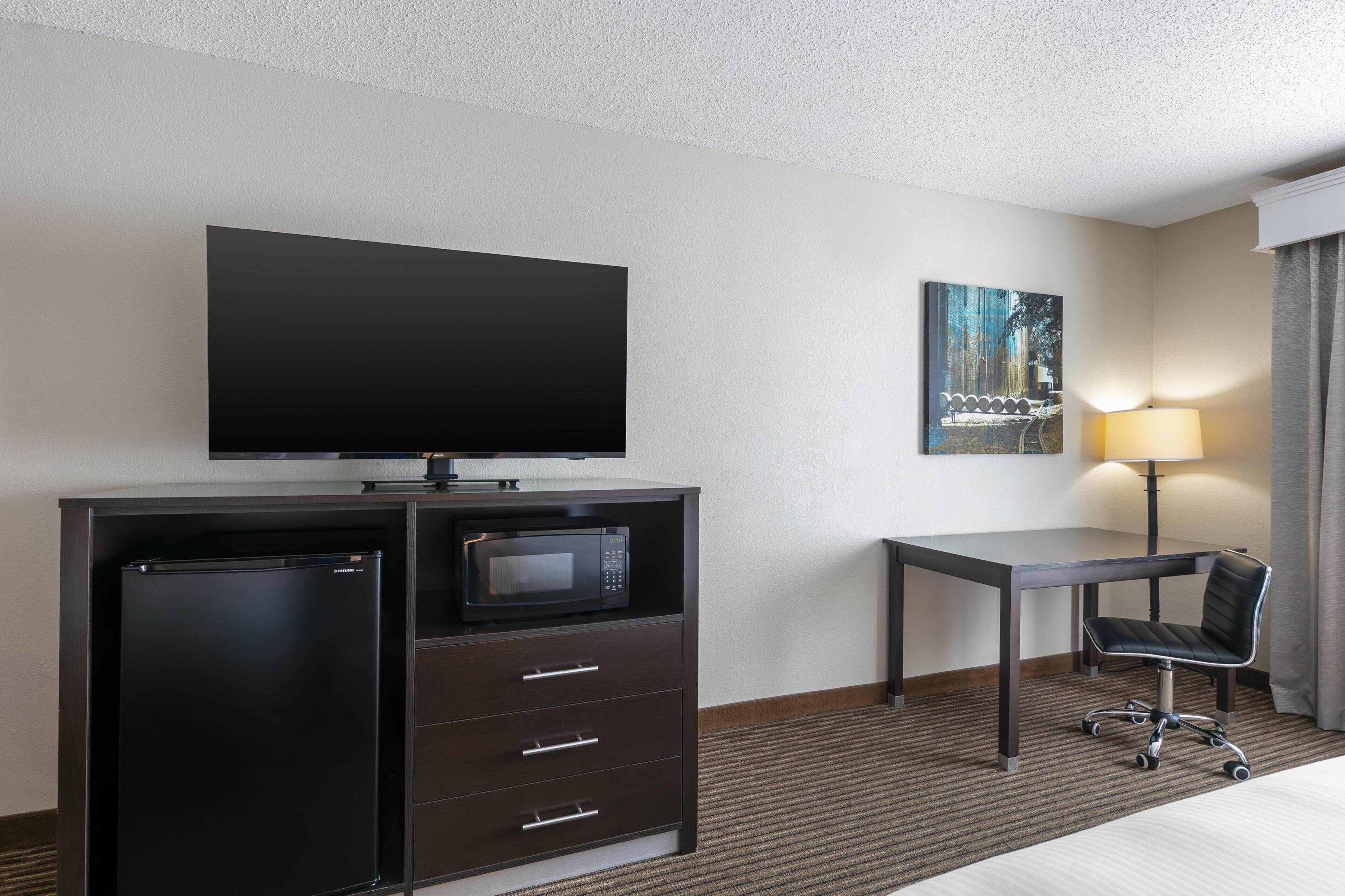 A hotel room with a flat-screen TV, a mini-fridge, a microwave, a desk, a chair, and a lamp.