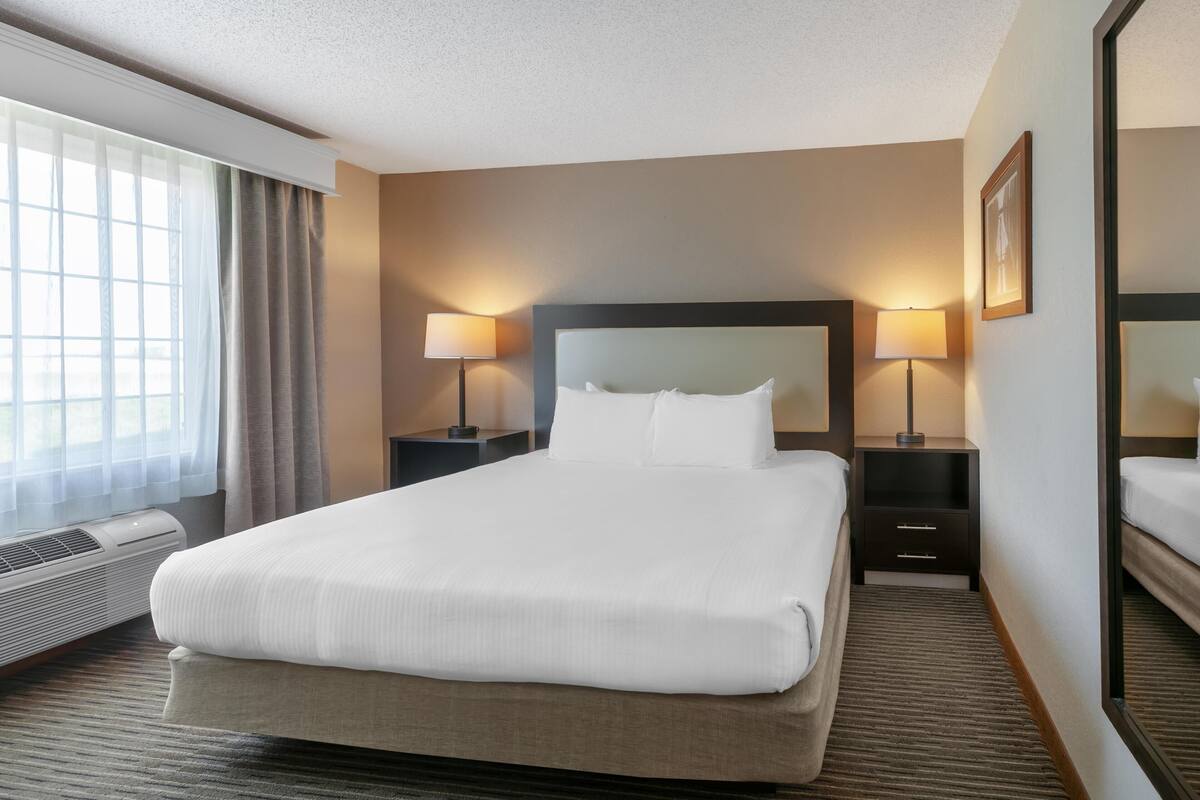 A hotel room with a large bed, two bedside tables with lamps, a window with curtains, and a wall-mounted mirror.