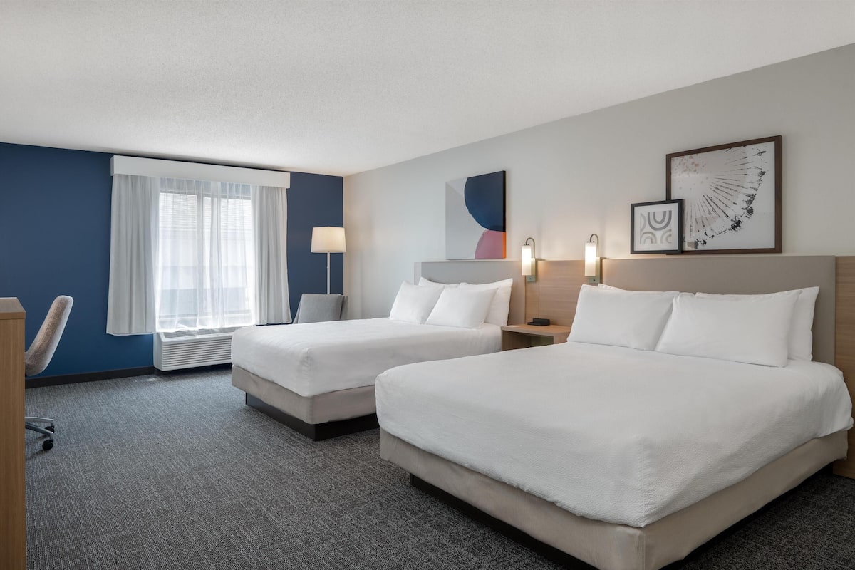Une chambre d’hôtel avec deux lits, un bureau, une chaise, une fenêtre avec des rideaux légers et des œuvres d’art murales.