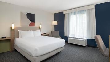 A hotel room with a large bed, a desk, a chair, a wall-mounted lamp, and a window with curtains.