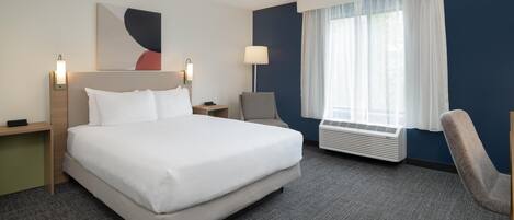 A hotel room with a large bed, a desk, a chair, a wall-mounted lamp, and a window with curtains.