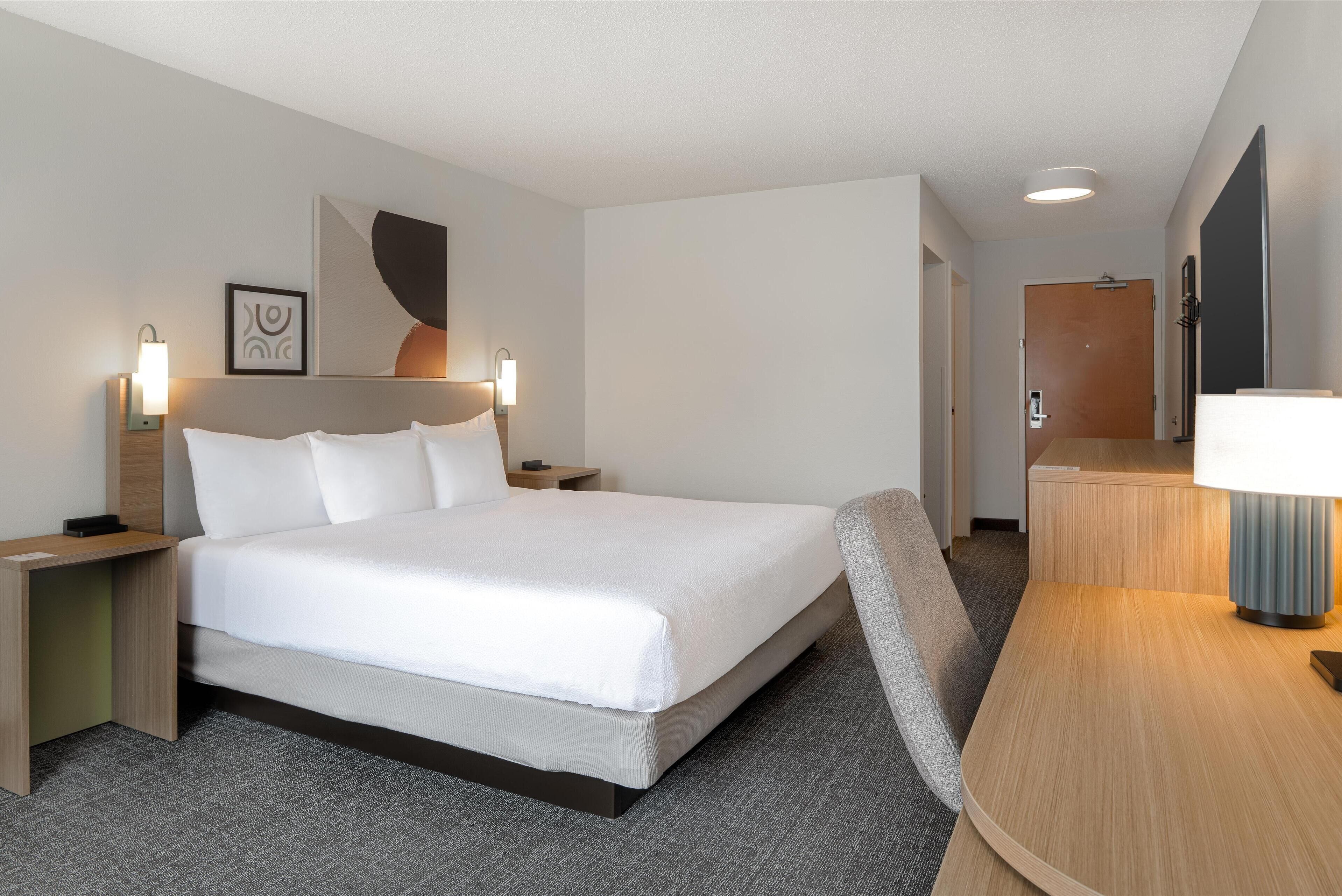 A hotel room with a large bed, bedside tables with lamps, a desk with a chair, and a door leading to another room.